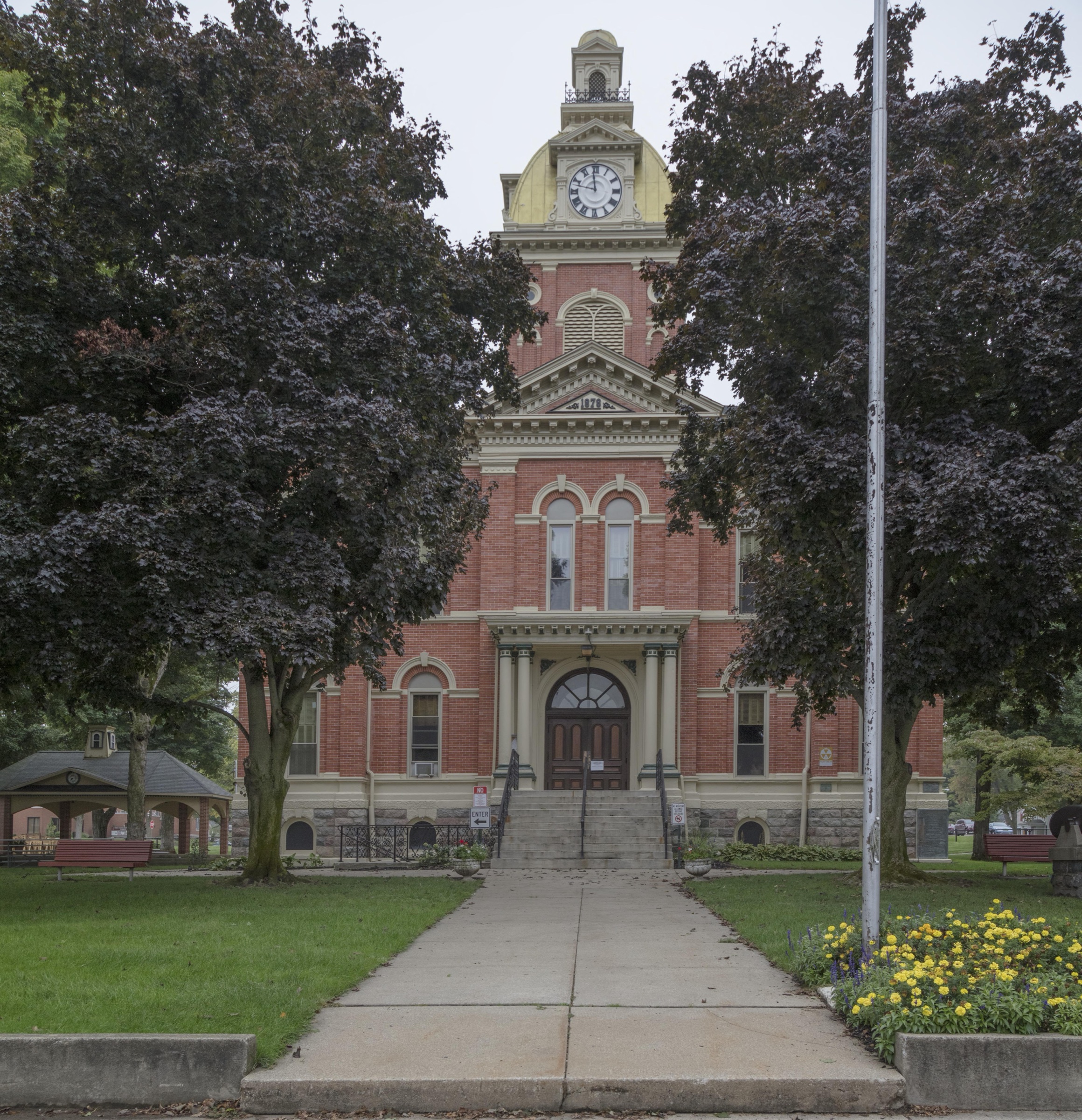 The LaGrange County Courthouse seen from the front walk, a red brick building with cream stone trim and a clock tower topped by a yellow dome, framed by two dark leaved maples, with a concrete walk running up to the arched entrance and marigolds along the edge of the lawn.