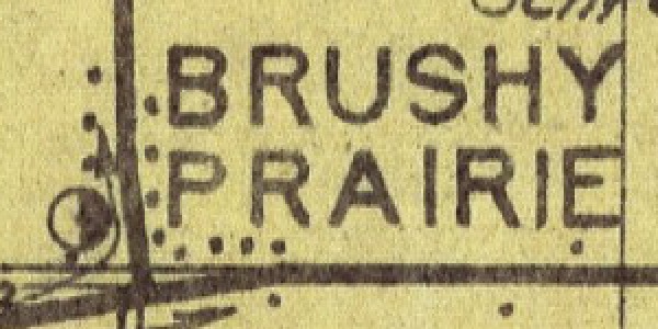 A small area of the map. The words BRUSHY PRAIRIE in capitals, and beside them, at a road corner, a small circle with its right half filled.